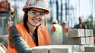 Lächelnde Bauarbeiterin mit Schutzhelm, Schutzbrille und orangefarbener Warnweste arbeitet an einer Mauer aus hellen Steinen.