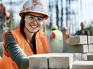 Lächelnde Bauarbeiterin mit Schutzhelm, Schutzbrille und orangefarbener Warnweste arbeitet an einer Mauer aus hellen Steinen.