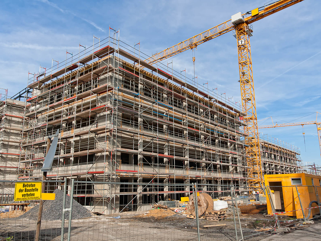 Mehrstöckiges Gebäude im Rohbau mit Baugerüst und mehreren Kränen auf einer weitläufigen Baustelle.