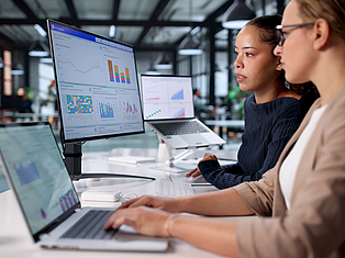 Zwei Frauen arbeiten an Laptops und betrachten Datenanalysen mit Diagrammen auf mehreren Bildschirmen.